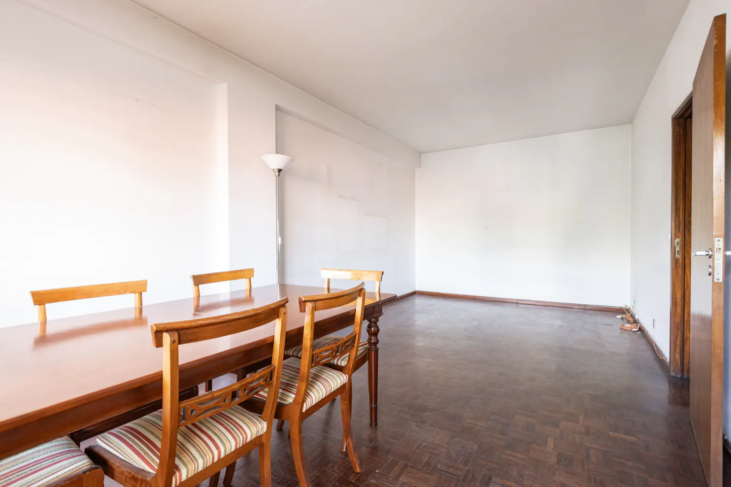 A dining room with a wooden table, chairs, and dark parquet flooring. White walls and an open wooden door are visible.