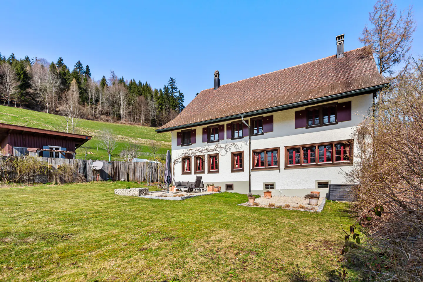 White two-story house with brown shutters and a tiled roof, set in a grassy yard against a backdrop of forested hills.