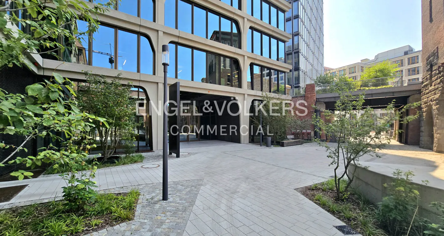Modern office building with glass facade and stone courtyard featuring small trees and a paved walkway.