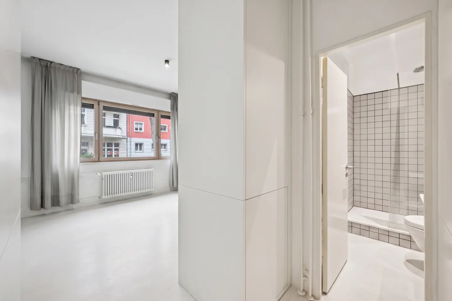 A bright, modern apartment interior with white walls, a window with gray curtains, and an open bathroom door revealing a tiled shower.