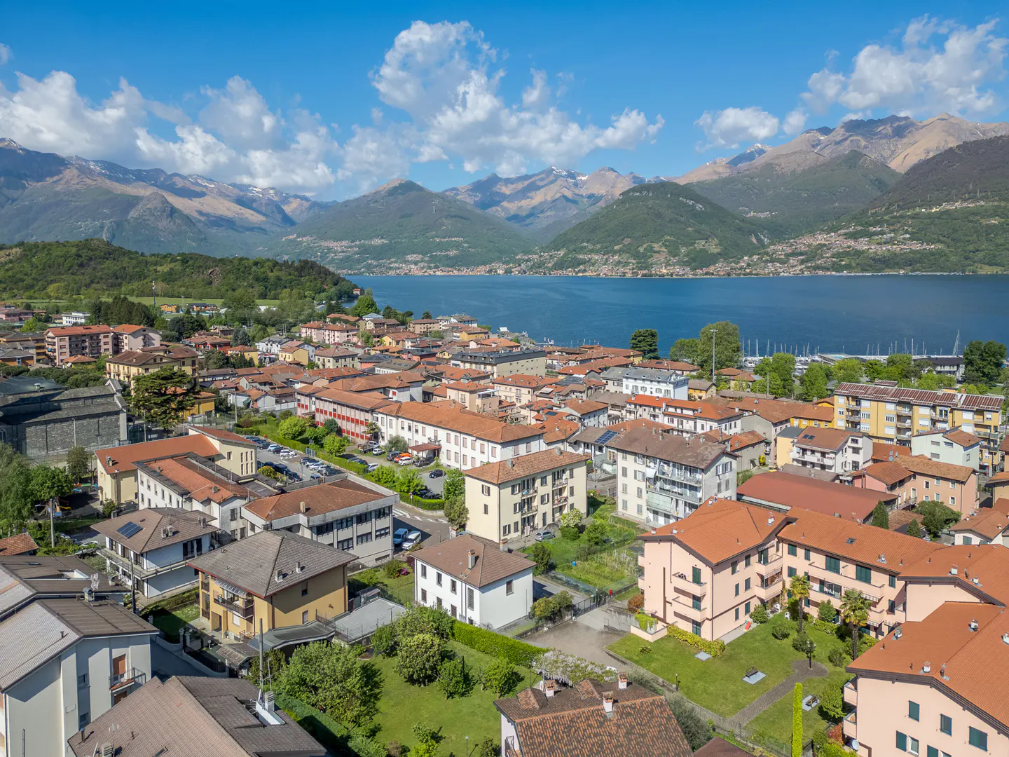 Aerial view of a town with red-tiled roofs, a blue lake, and mountains in the background under a partly cloudy sky.