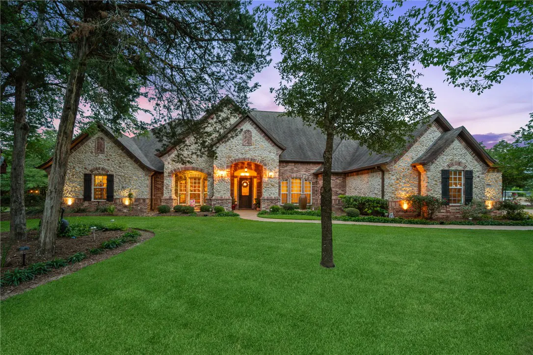 Front view of a one-story stone and brick house with a green lawn at dusk. The house has a dark roof and arched entryway.