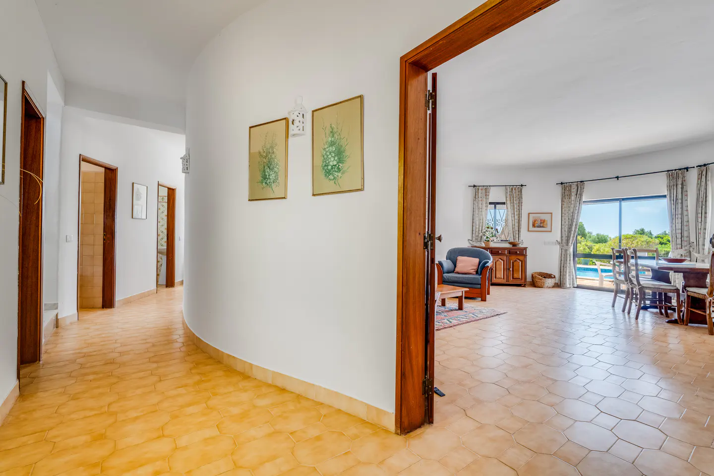 Hallway with tan tile floor and white walls. A living room with a blue chair and pool view is visible through a wood door frame.