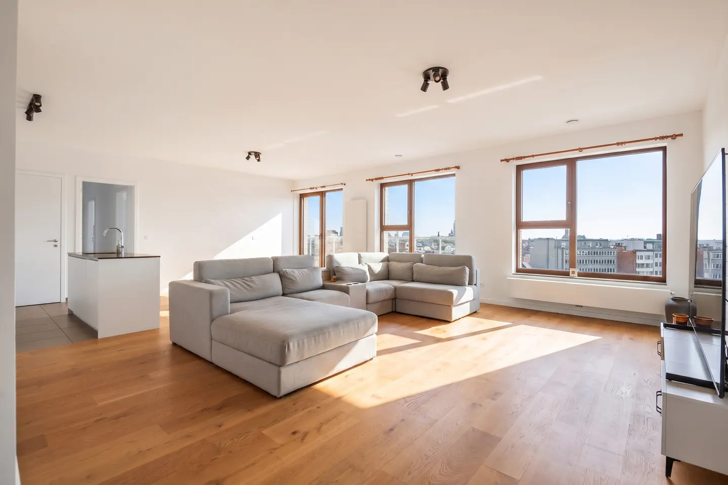 Bright, modern living room with a gray sectional sofa, wood floors, and large windows overlooking a city. White kitchen island visible.
