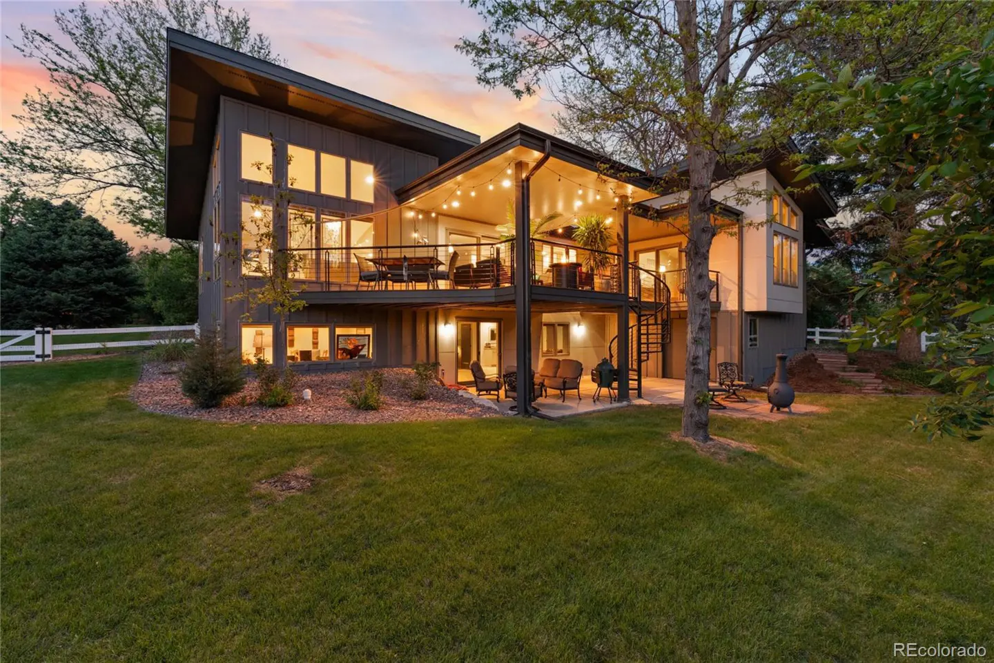Two-story gray house with large windows and a deck with string lights, viewed from the lawn. A spiral staircase leads to the patio.