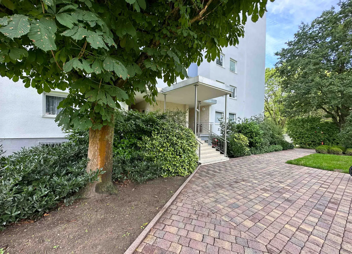 Exterior view of a white apartment building with a brick driveway and green trees.