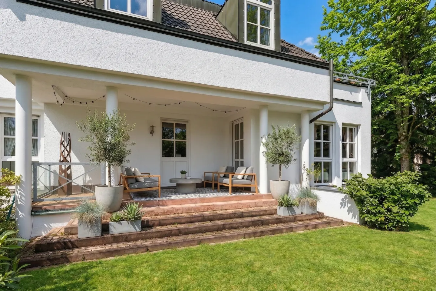 Exterior view of a white house with a covered patio, featuring outdoor seating, potted plants, and a green lawn.
