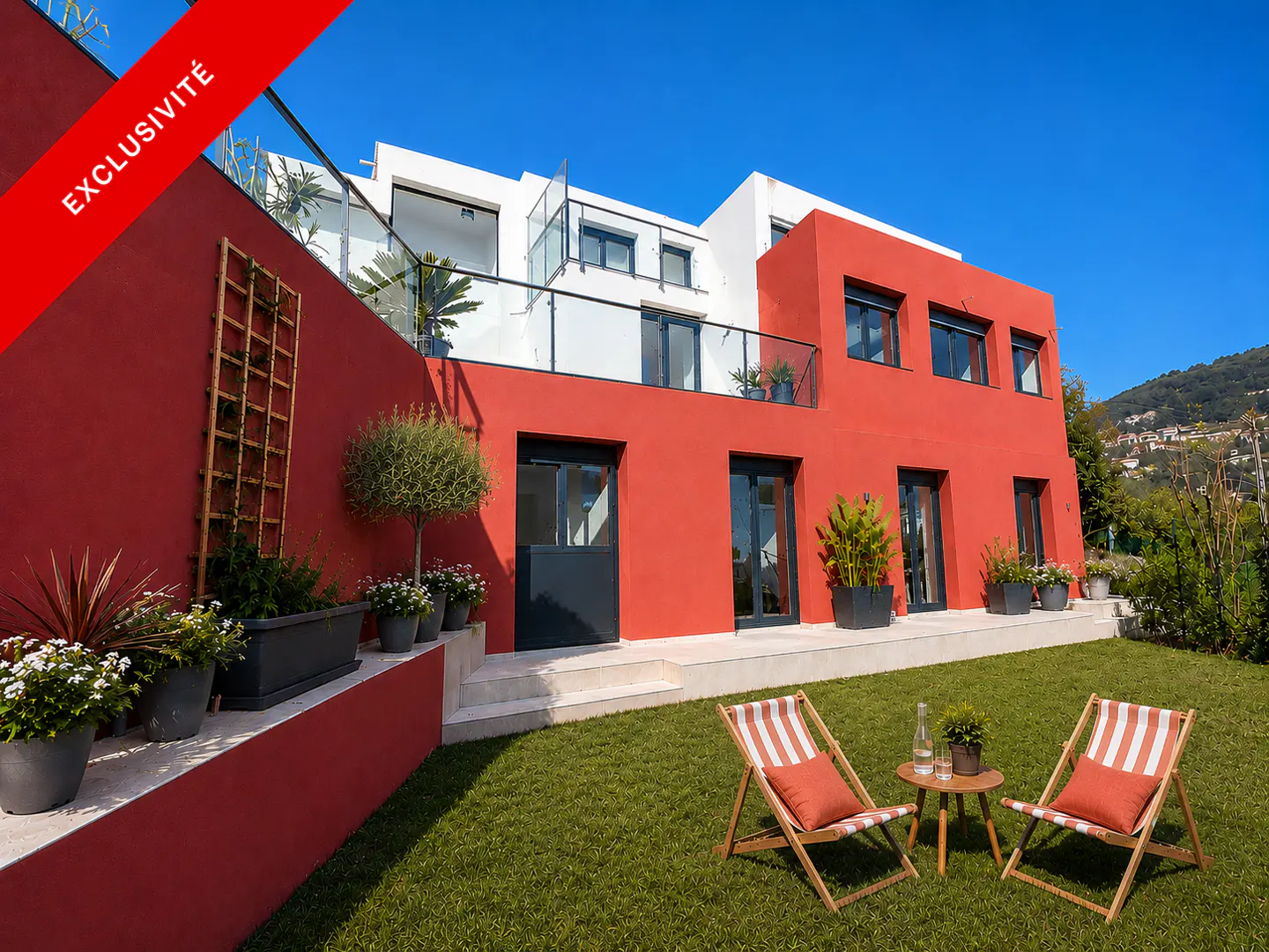 Modern red and white house with a green lawn and two striped deck chairs. A small table sits between the chairs with a water bottle.
