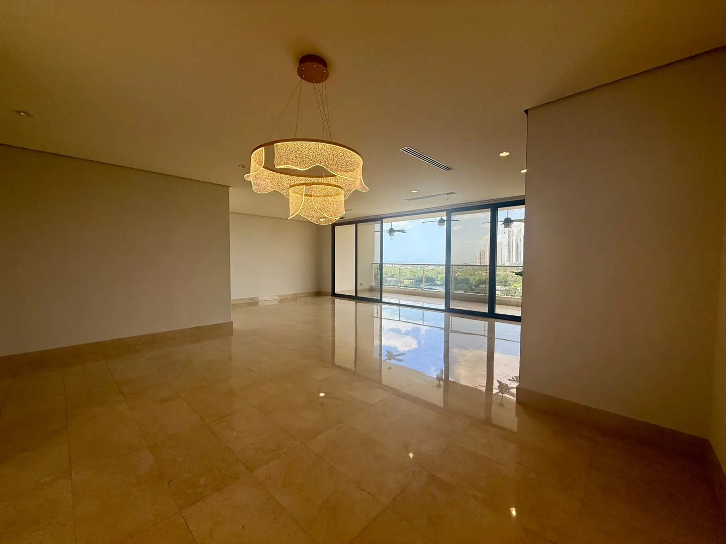 Spacious, empty room with marble floors and a modern chandelier. Large sliding glass doors lead to a balcony with a view of the ocean and city.