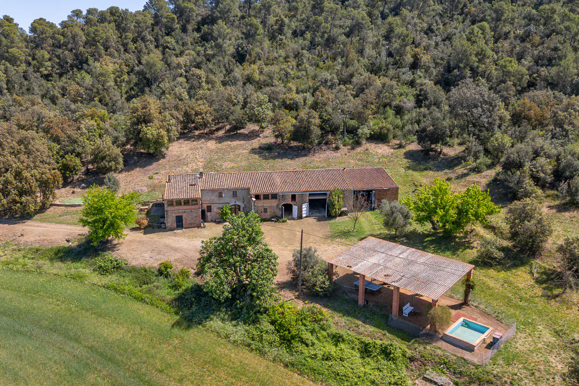 Aerial view of a rural property with a long, red-tiled roof house, a covered patio, and a small pool, surrounded by green fields and a dense forest.