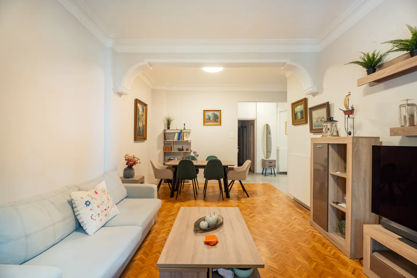 A bright living room with a light blue sofa, wooden coffee table, and dining area with a wooden table and chairs. The floor is parquet.