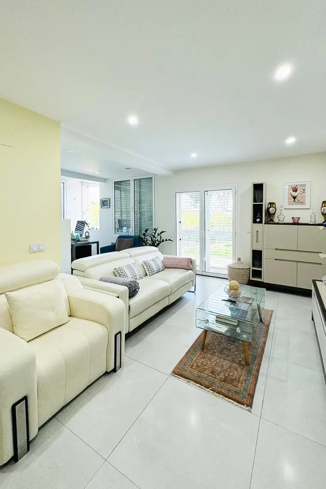 Bright living room with white tile floors, cream sofas, glass coffee table on a rug, and sliding glass doors to the outside.