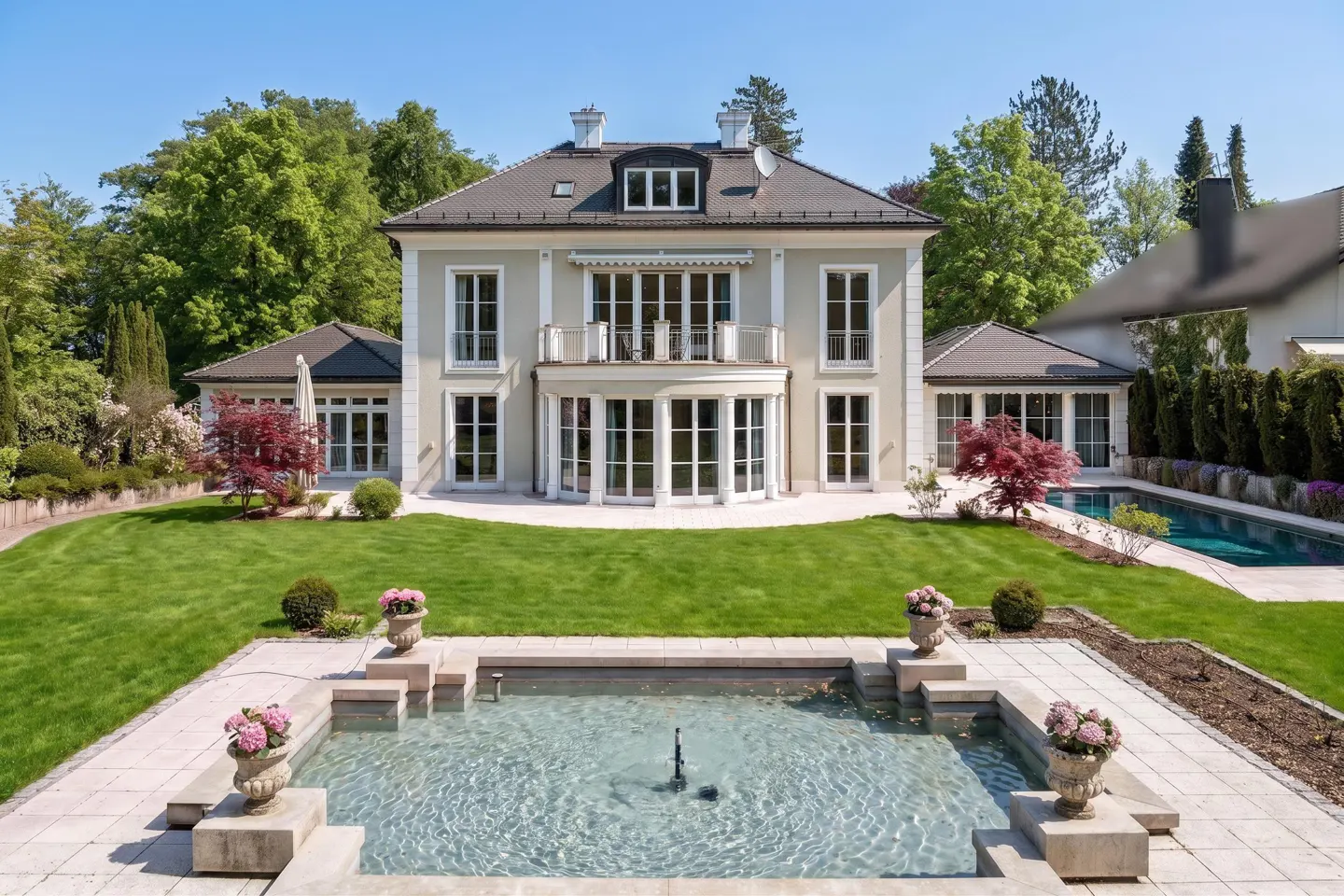 A grand beige mansion with a pool, fountain, and lush green lawn under a clear blue sky.