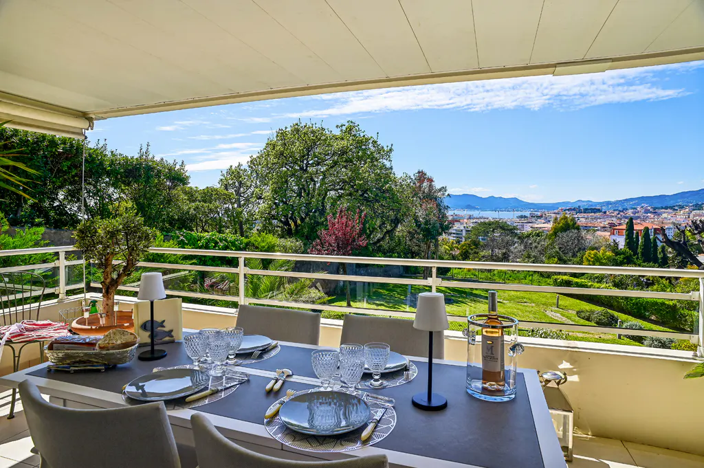 Outdoor dining area on a balcony with a table set for six, overlooking a green landscape and cityscape under a blue sky.