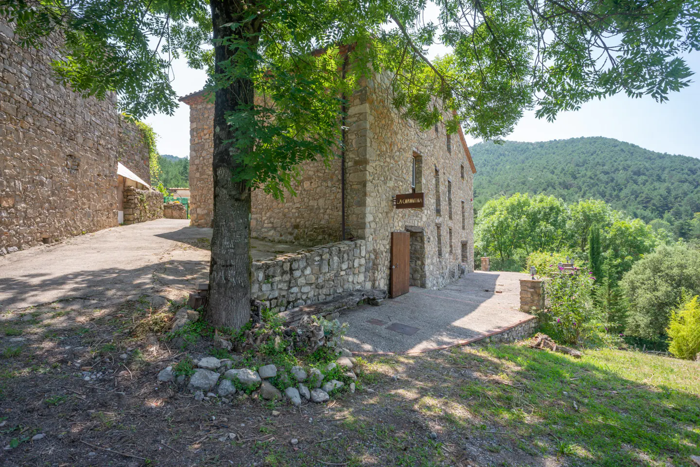 Exterior view of a stone house with a brown door and a sign, surrounded by trees and a mountain in the background.