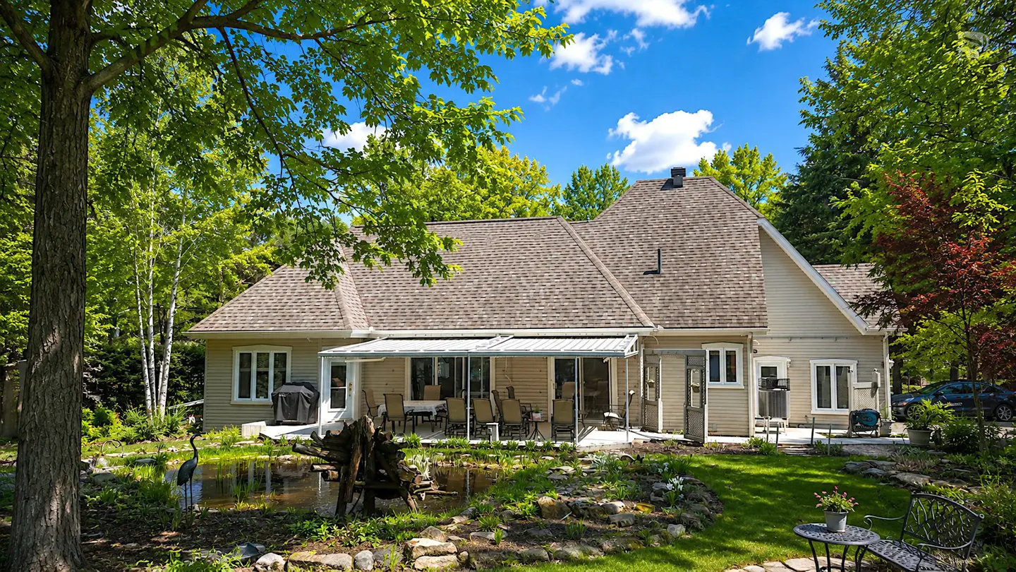 Backyard view of a beige house with a brown roof, a pond, and a patio with a table and chairs. Green trees surround the house.