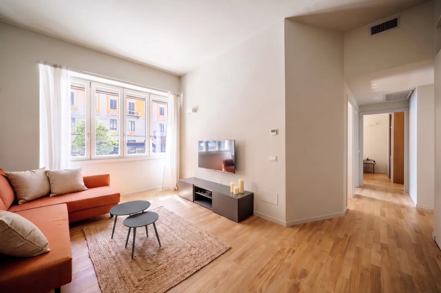 Bright living room with orange sofa, two-tiered table, and TV. Large window with white curtains overlooks a city street. Hardwood floors and neutral walls.