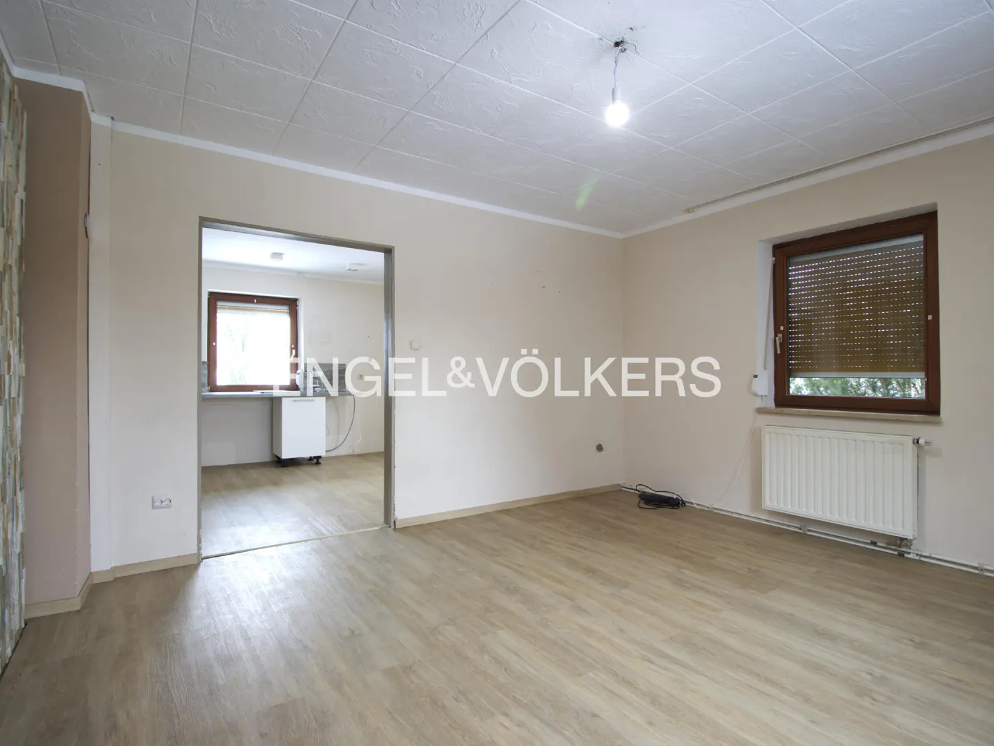 A bright, empty room with light wood floors, beige walls, and a window with a brown frame. A doorway leads to a kitchen with white cabinets.