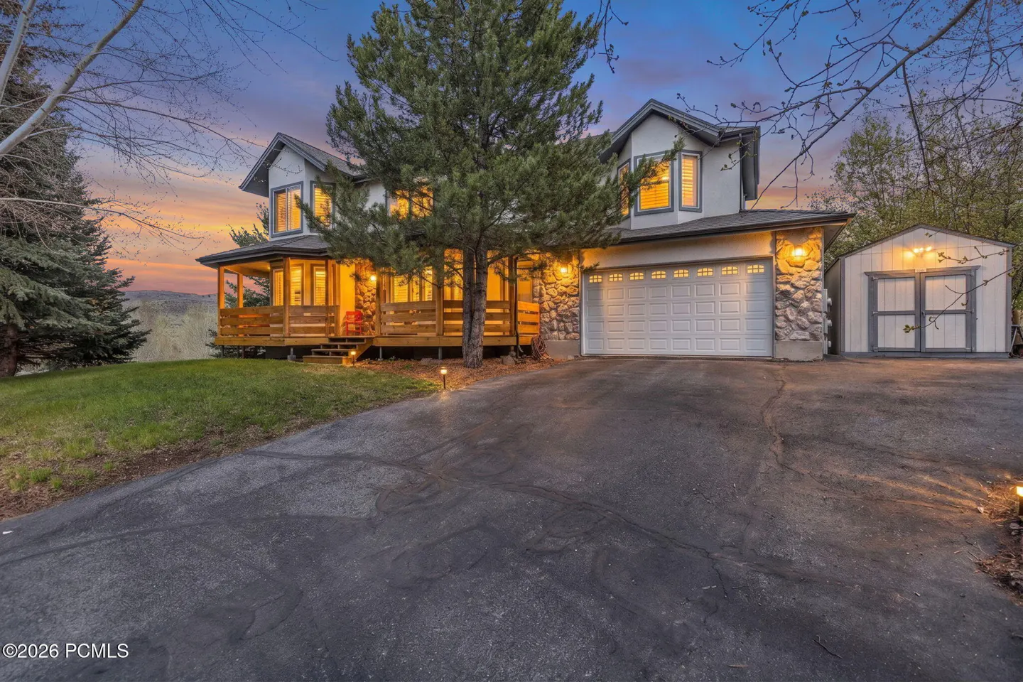 Two-story house with a wooden porch, stone accents, and a white garage door. A shed is visible on the right. The sky is a mix of blue and orange.