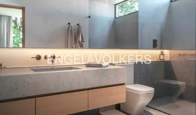 Modern bathroom with a gray stone countertop, light wood cabinets, and a white toilet. A large mirror reflects the room.