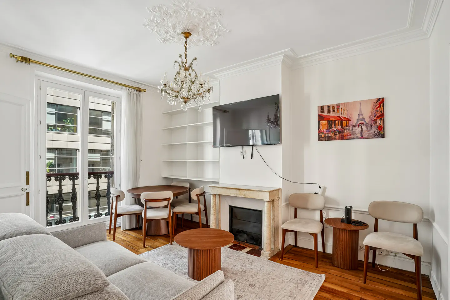 Bright living room with a gray sofa, round wood tables, and a dining set. A chandelier hangs from the ceiling, and a Paris painting adorns the wall.