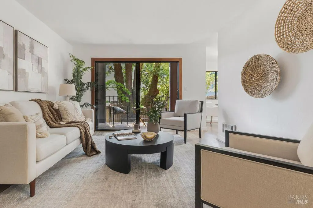 Living room with white sofa, black round table, and balcony view of trees. Woven baskets hang on the wall.