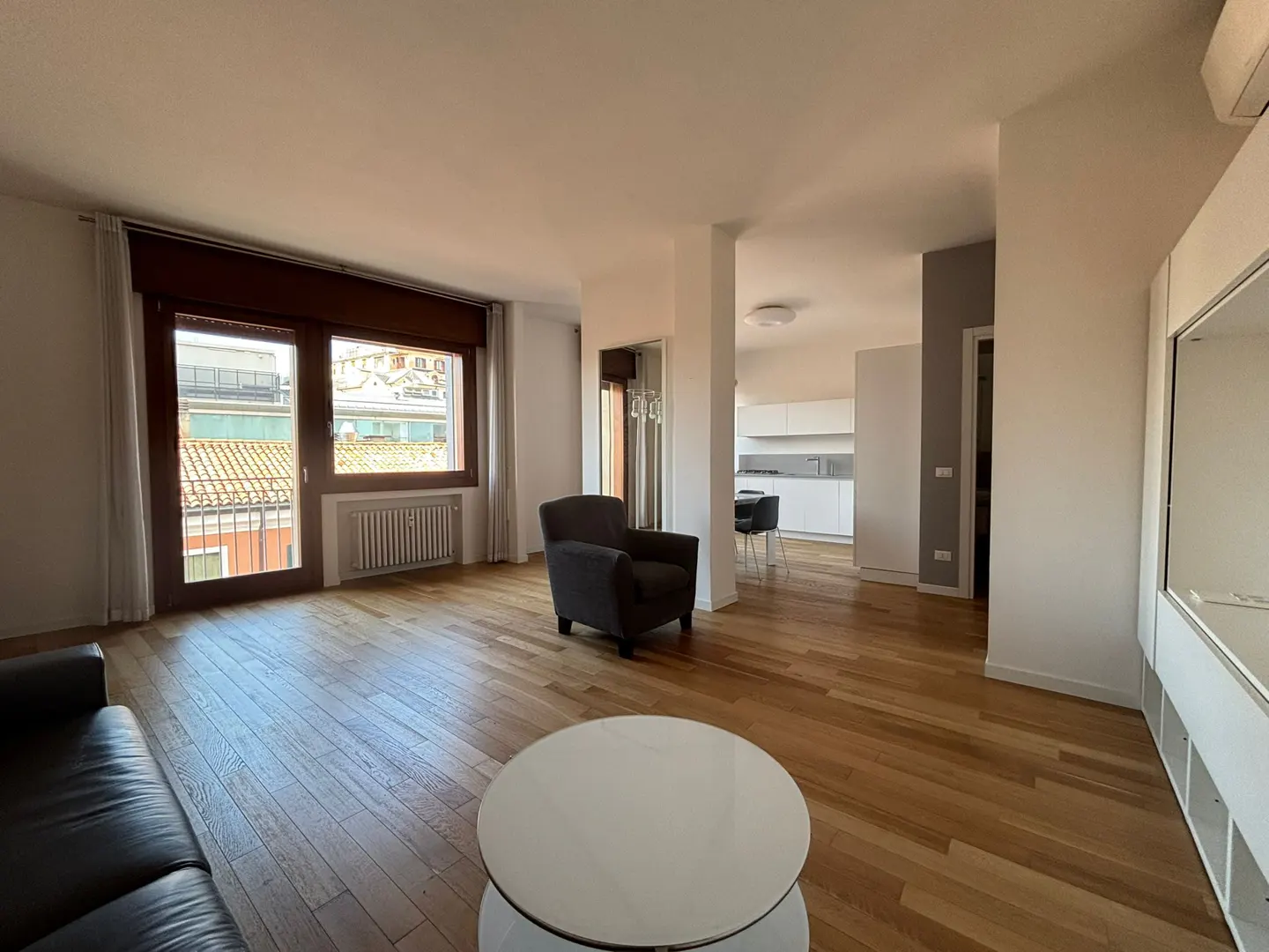 Bright living room with wood floors, a gray armchair, and a white coffee table. A window overlooks a city view. The kitchen is visible in the background.