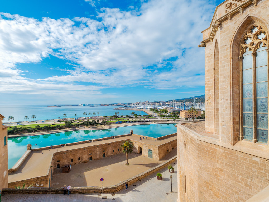 View of a stone cathedral wall overlooking a turquoise reflecting pool, a historic fortress, and the Mediterranean sea.