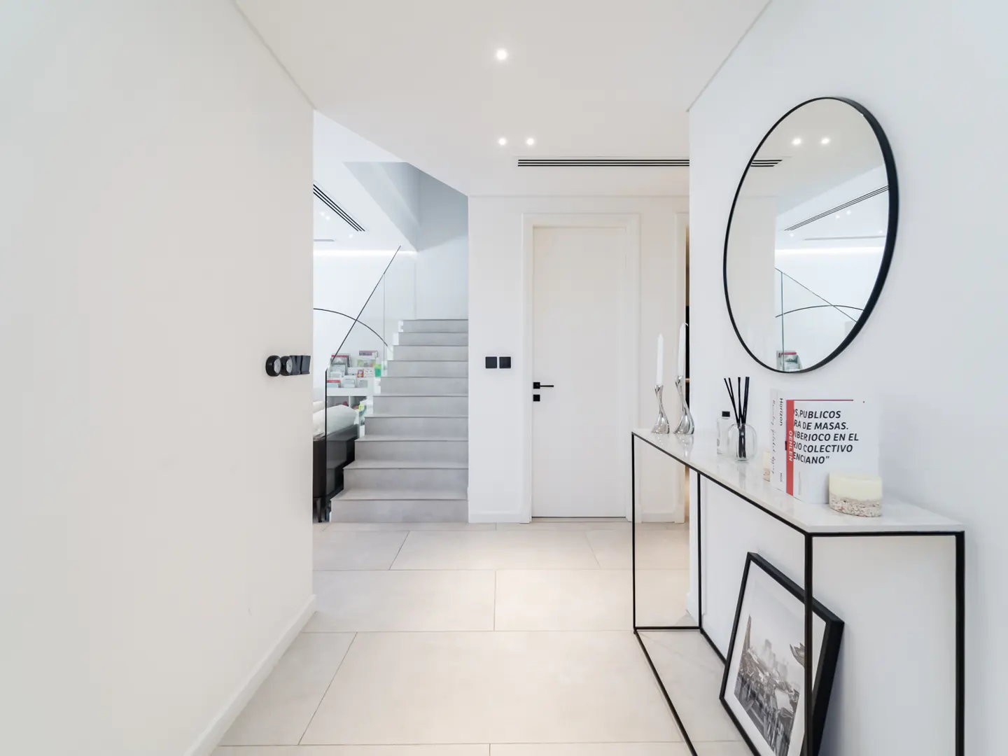 Bright, modern entryway with white walls, tile floor, and stairs. A black-framed mirror hangs above a console table with decor.