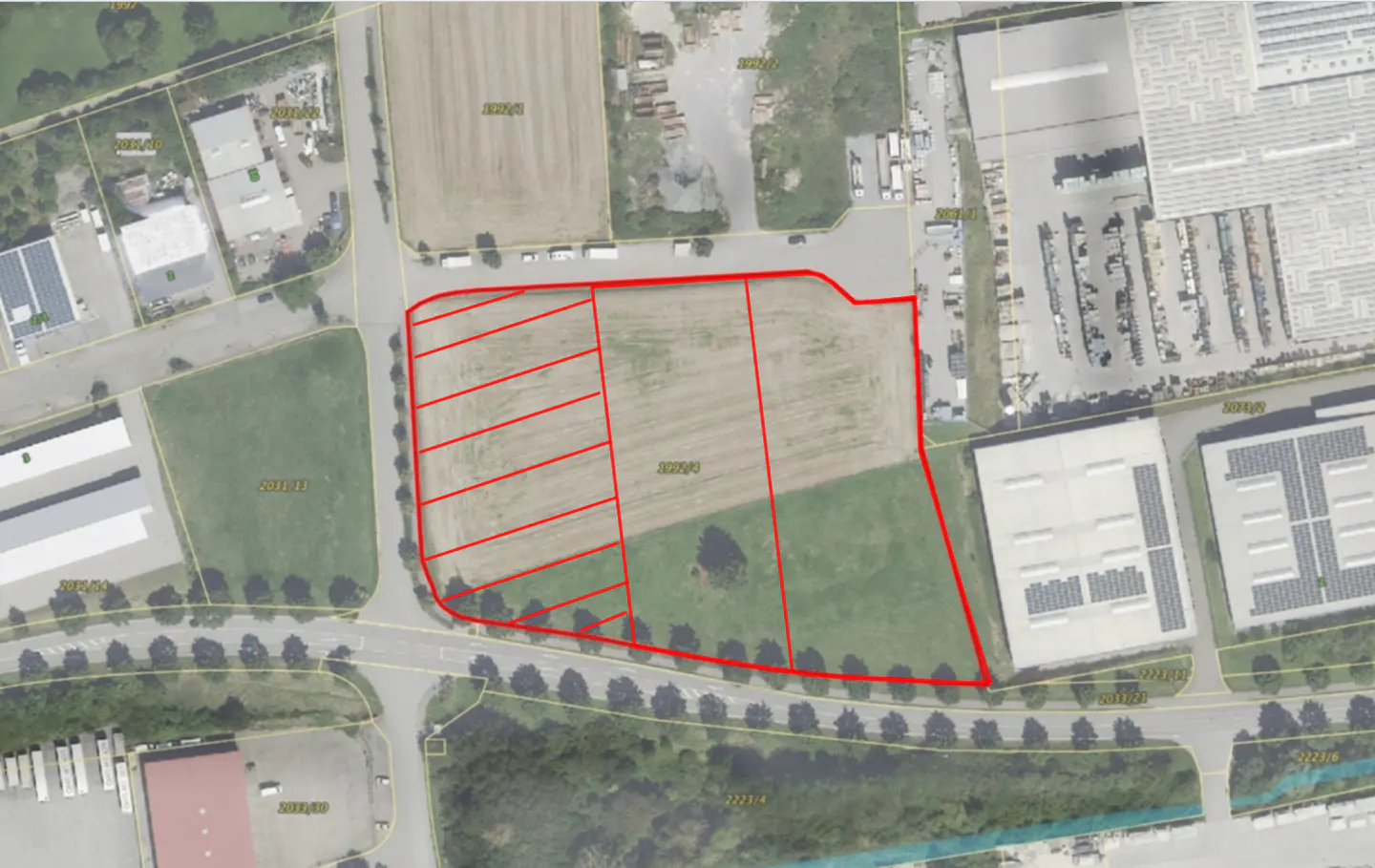 Aerial view of a property outlined in red, divided into smaller plots, surrounded by roads, trees, and industrial buildings.