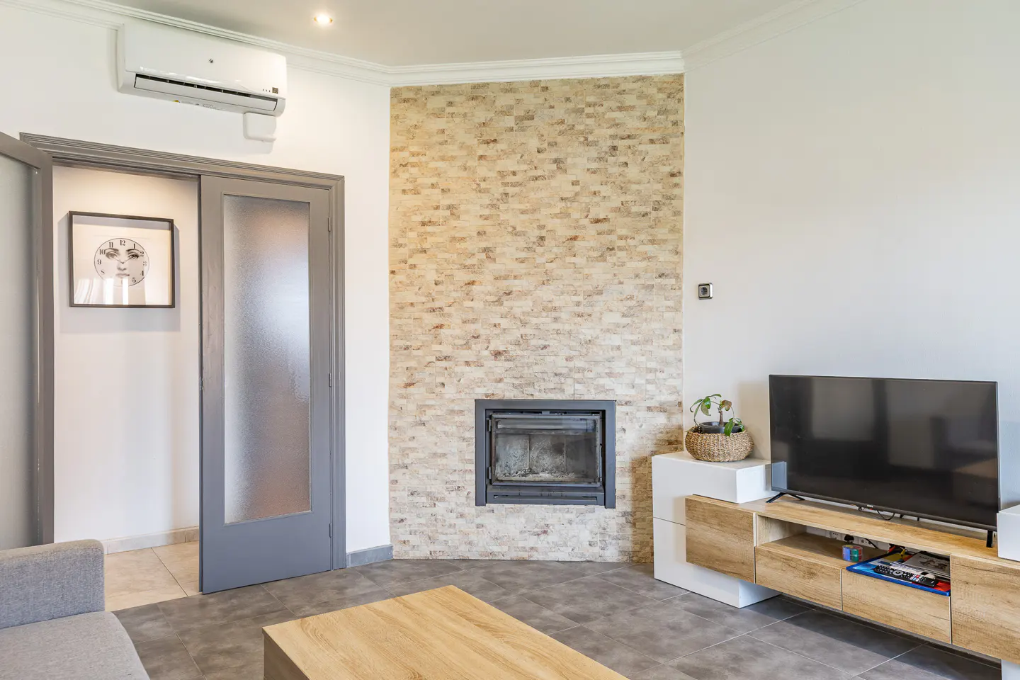 Living room with a stone fireplace, gray door, and a TV on a wooden stand. An air conditioner is mounted on the wall.
