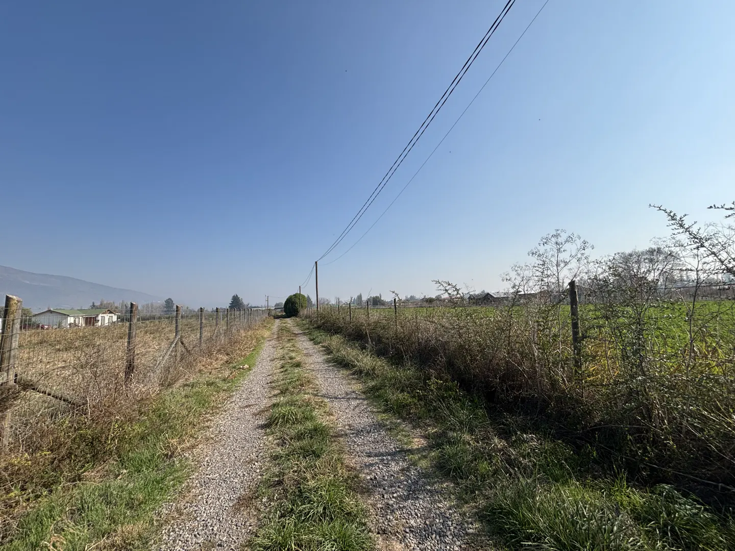 A gravel path leads through a rural landscape with wire fences, dry grass, and power lines under a clear blue sky.