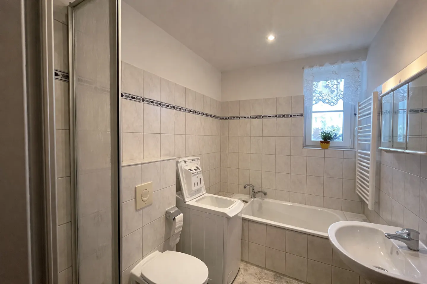 Bright bathroom with beige tile, a shower, toilet, sink, and bathtub. A small window with a plant sits above the tub.