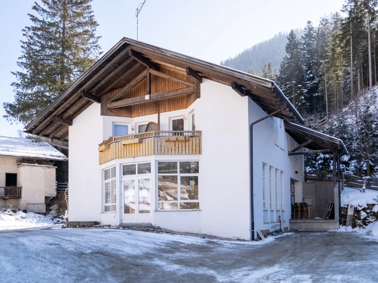 A two-story white building with a wooden balcony and roof, set against a snowy, wooded hillside.