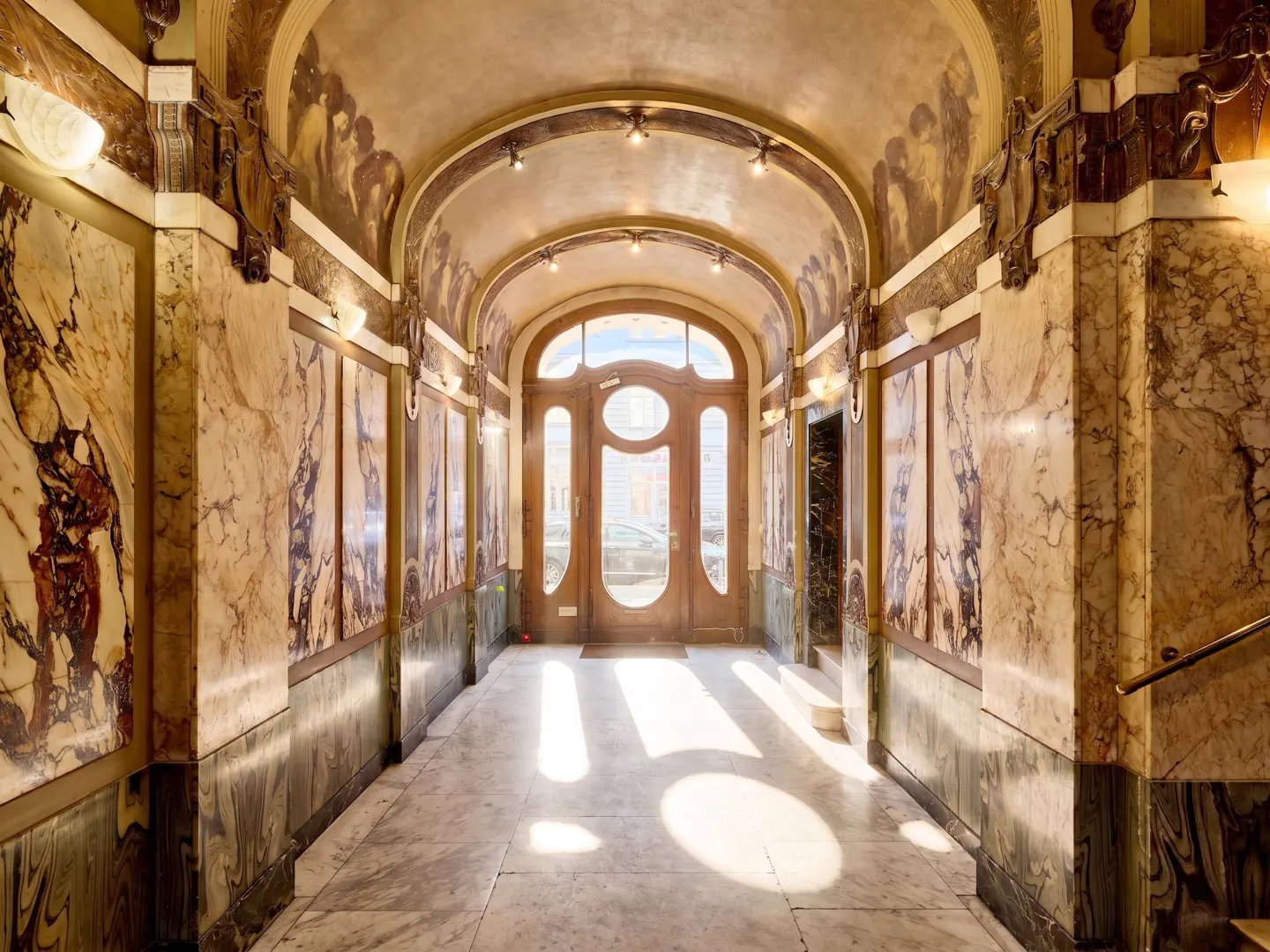 Arched hallway with marble walls and floor leads to a wooden door with glass panels. Light shines through the door.