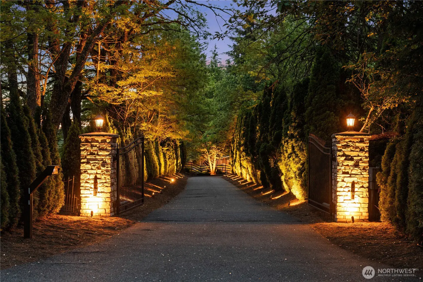 Gated driveway entrance with stone pillars and lanterns, lined with trees and shrubs, illuminated at dusk.