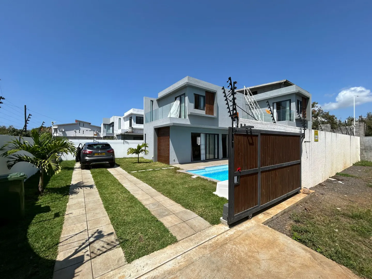 Exterior of a modern two-story gray house with a pool, viewed from the driveway through an open brown gate. A car is parked on the driveway.