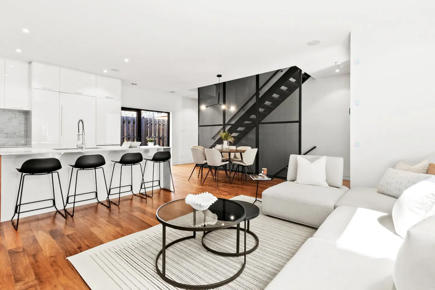 Bright, open-concept living space with white walls, wood floors, and modern furnishings. Kitchen island with black stools, dining table, and staircase visible.