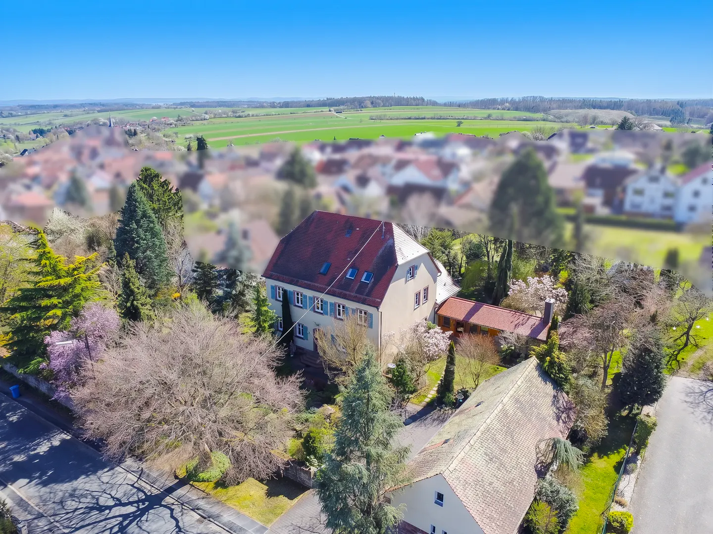 Aerial view of a beige two-story house with a red roof and blue shutters, surrounded by trees and a blurred village background.