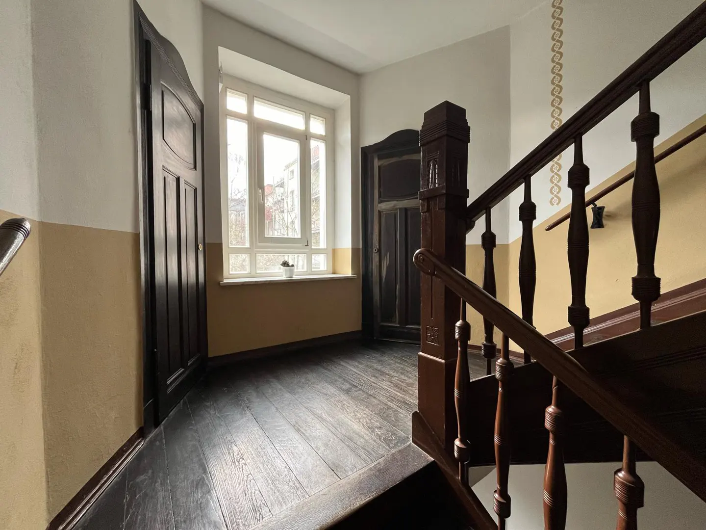 Hallway with dark wood floors, staircase with brown banister, and two dark wood doors. A window with white trim is in the background.