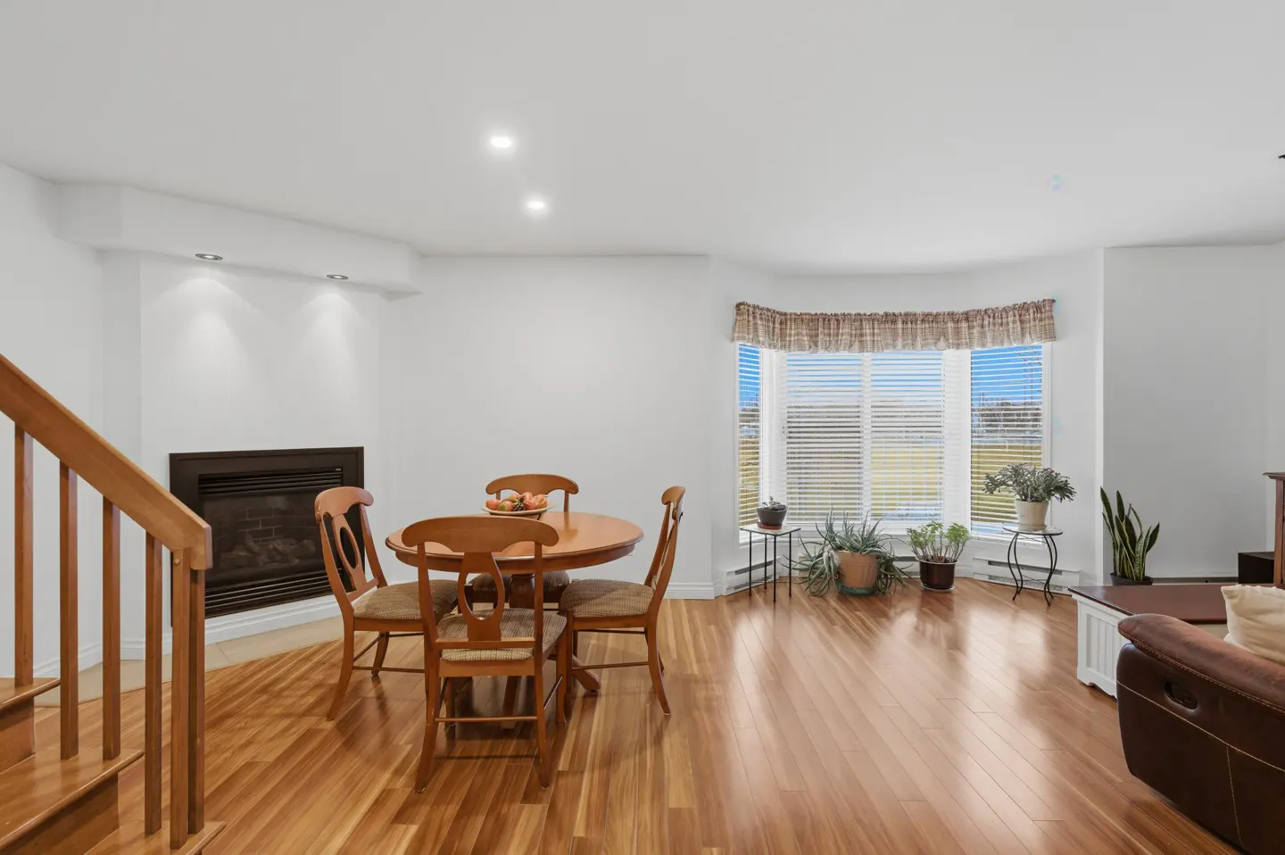 Bright living room with hardwood floors, a round wooden table with chairs, a fireplace, and a bay window with plants.