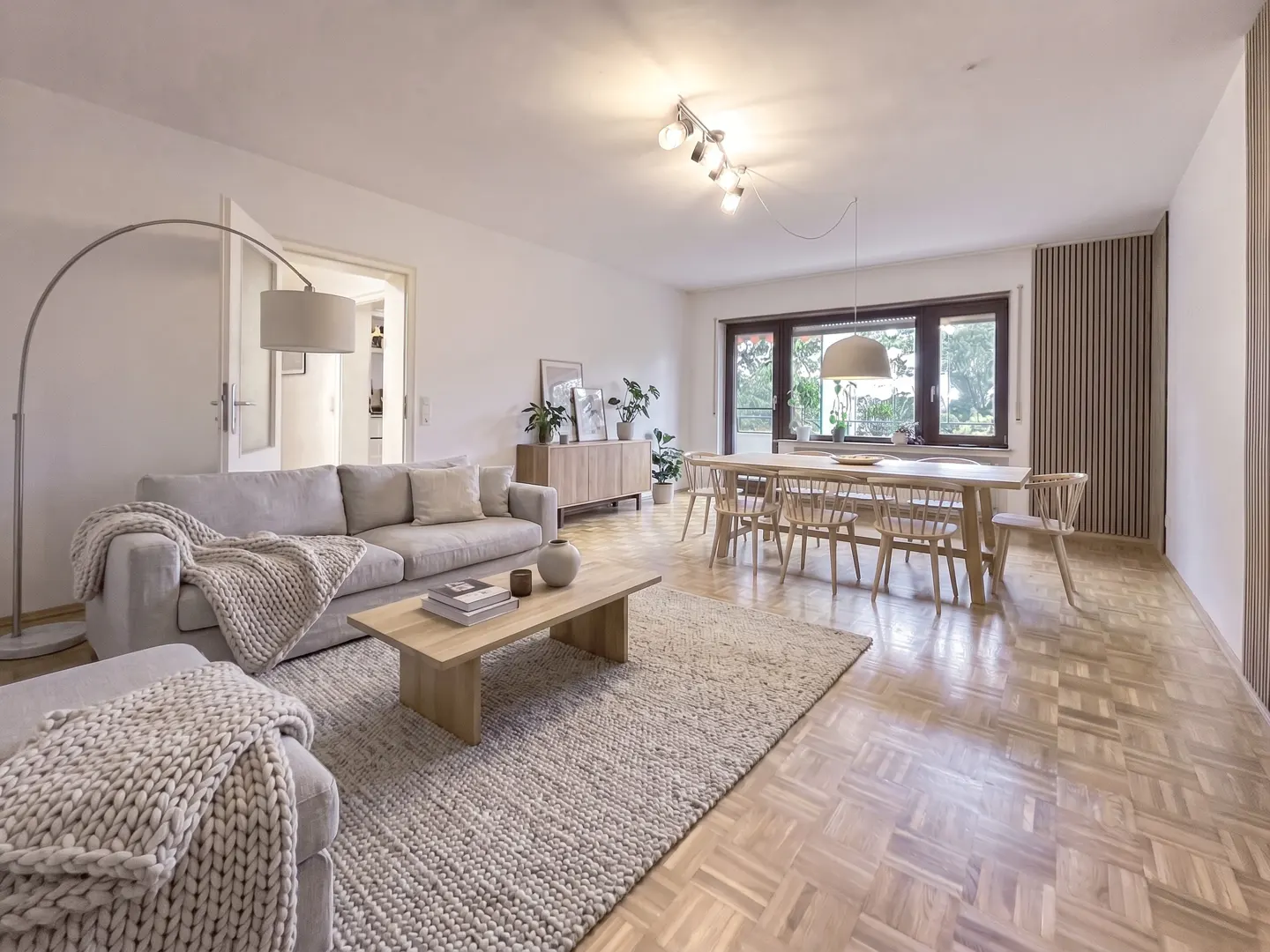 Bright, open-plan living and dining room with light wood floors, a grey sofa, coffee table, and wooden dining set.