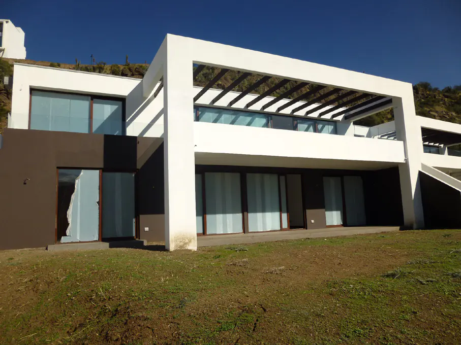 Modern two-story house with white frame and brown walls, set on a grassy hill under a blue sky. One window is broken.