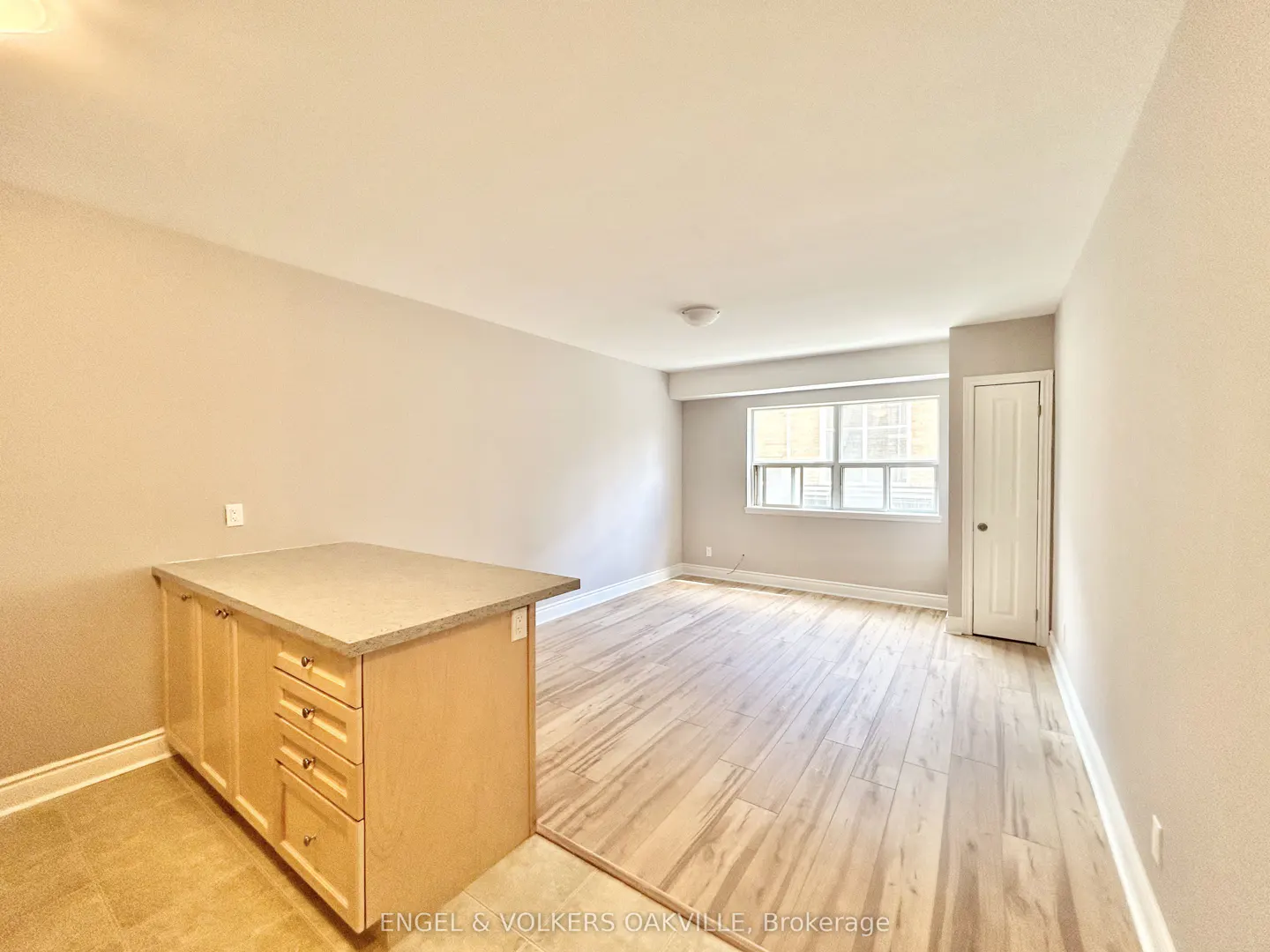 A bright, empty room with light wood floors, beige walls, a window, and a kitchen island.