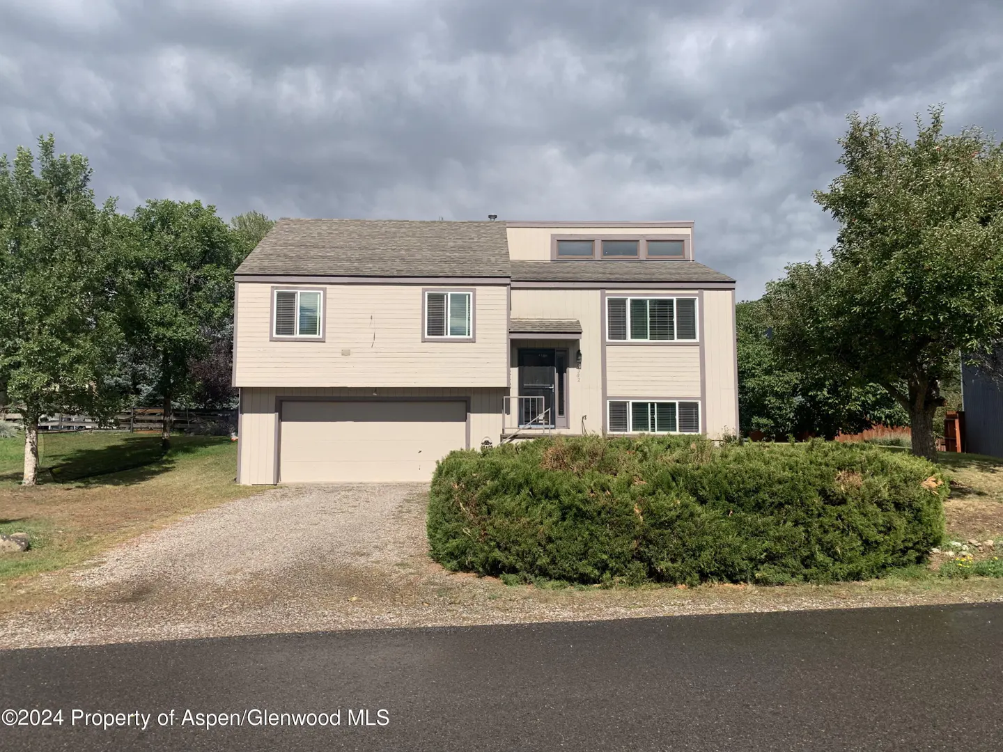 Two-story beige house with a gray roof, garage, and green bushes in front. Trees are visible in the background under a cloudy sky.