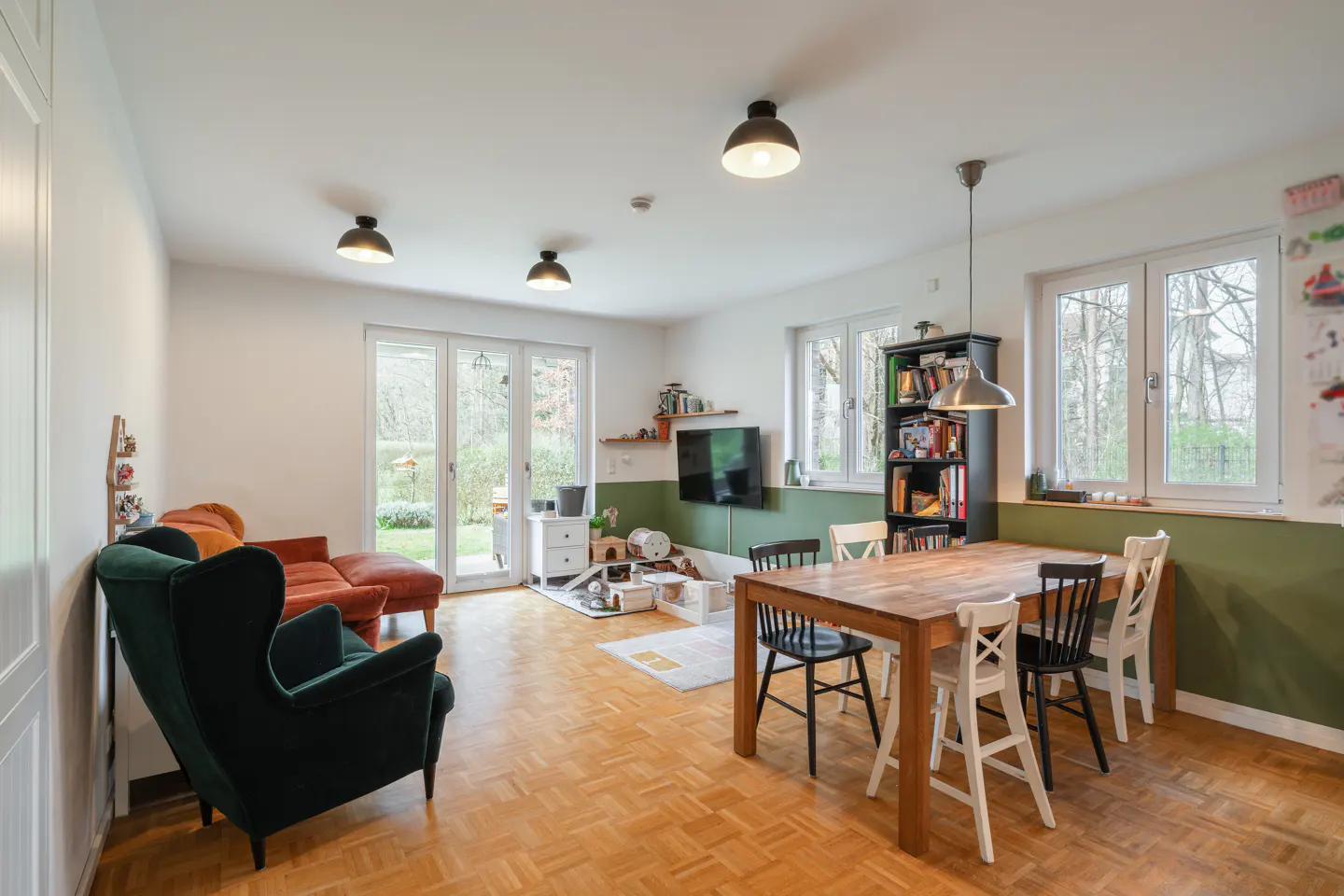 Bright living room with wood floors, green accent wall, and a wooden dining table with black and white chairs. A green armchair and orange sofa are visible.