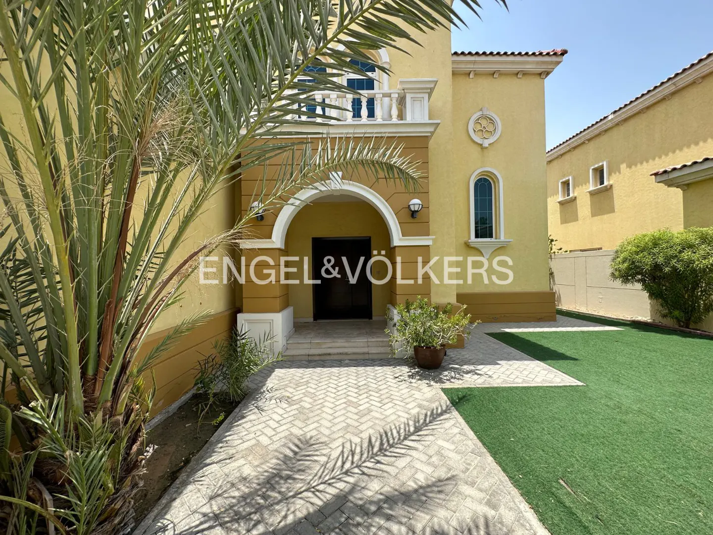 Yellow villa entrance with a paved walkway, palm tree, and green lawn under a clear blue sky.