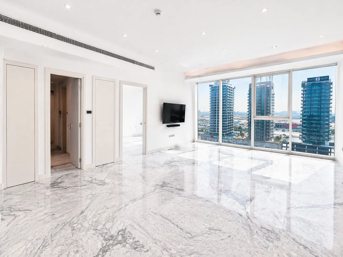 Bright, empty room with marble floors, white walls, and large windows showing city buildings. A TV is mounted on the wall.
