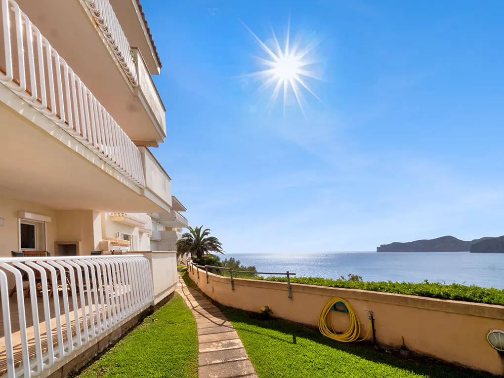 Exterior view of a beige apartment building with white railings, a stone path, and ocean views under a bright blue sky.