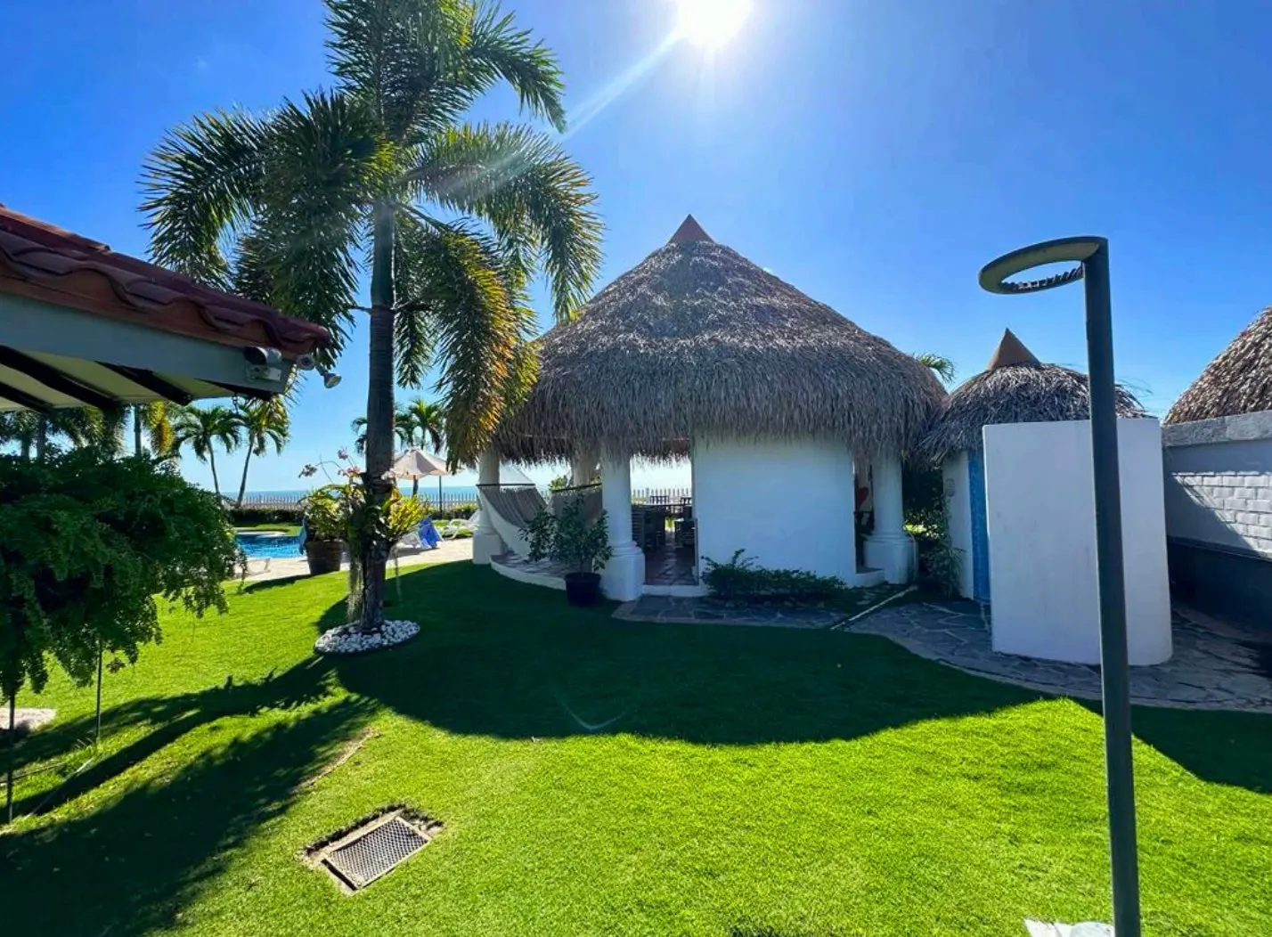 Lush green lawn with a white cabana featuring a thatched roof, palm tree, and blue sky.
